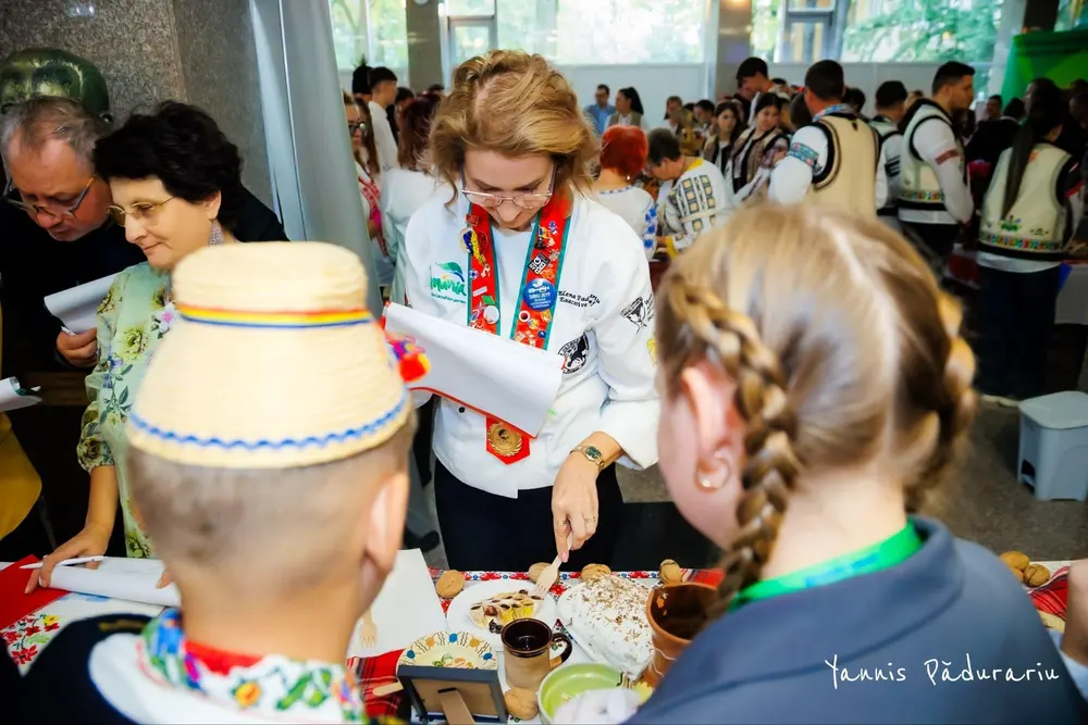 Chef Elena Pădurariu evaluează un preparat, cu doi concurenți în costume populare, fotografiați din spate
