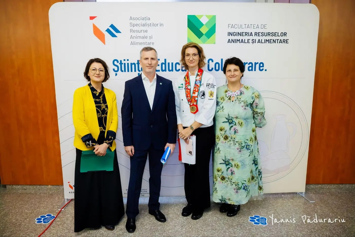 Chef Elena Pădurariu, în uniformă de bucătar, alături de organizatori, în fața panoului Facultății de Ingineria Resurselor Animale și Alimentare